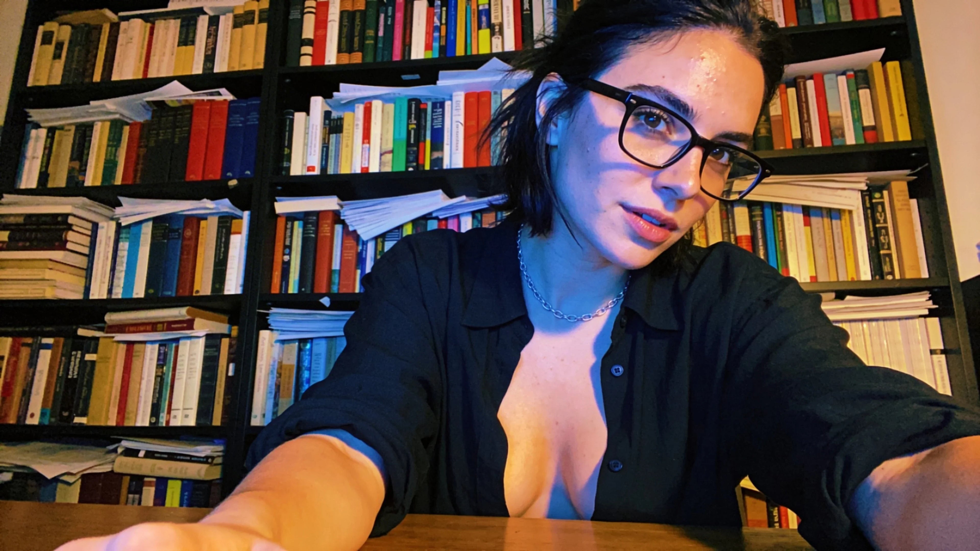 Woman with glasses and dark hair sitting at a desk in front of a bookshelf filled with books.