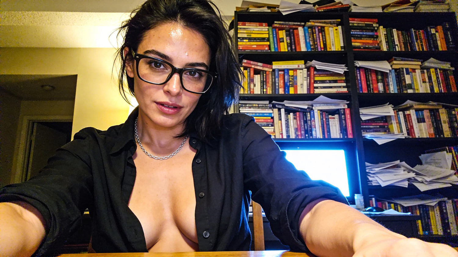 Woman with dark hair and glasses in black shirt, sitting at desk in front of bookshelf